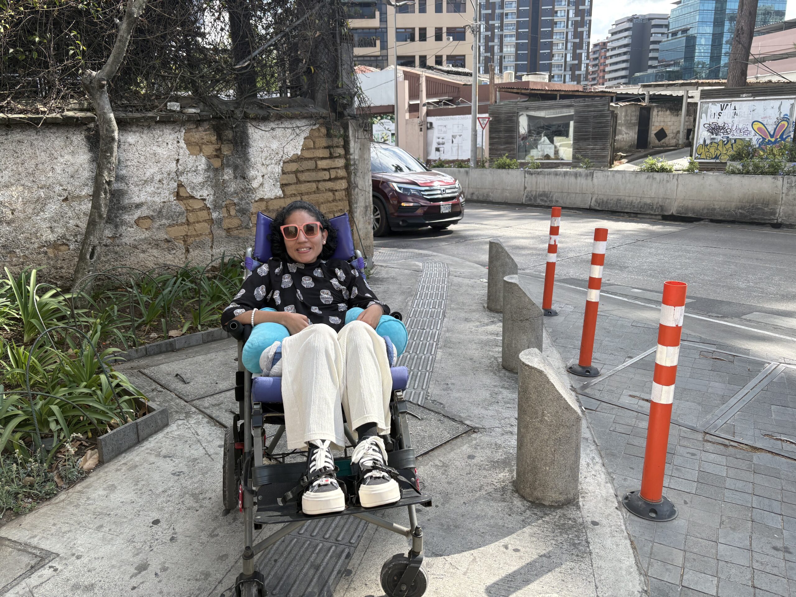 Una fotografía horizontal. A la izquierda, plantas y una pared antigua con ladrillo y un poco de pintura blanca. En medio. Andrea usuaria de silla de ruedas con un pantalón color beige y una blusa negra con búhos blancos está sobre una banqueta. A la derecha, obstáculos cilíndricos de cemento y conos anaranjados. Al fondo edificios y automóviles, pasando por la calle.
