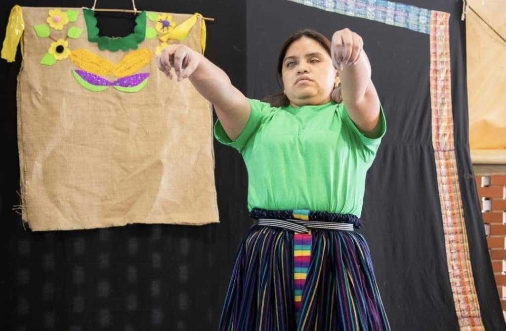 Isela está de pie frente a un fondo oscuro, vistiendo una camiseta verde y una falda larga de indumentaria maya. Tiene los brazos extendidos hacia adelante, con las manos inclinadas hacia abajo. A su lado, cuelga un huipil hecho de papel. 