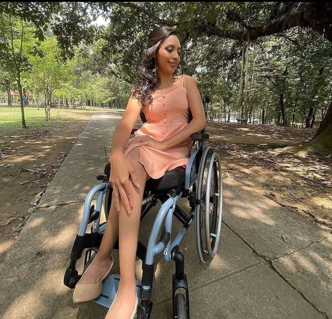 Una mujer joven está sentada en una silla de ruedas en medio de un camino de parque rodeado de árboles. Lleva un vestido color durazno y zapatos claros. Está sonriendo suavemente y mirando hacia un lado, con una postura relajada y elegante. La luz del sol atraviesa los árboles y crea un ambiente tranquilo y natural alrededor de ella. 