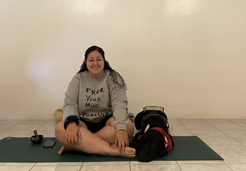 Marce está sentada sobre un tapete de yoga, con las piernas cruzadas y una postura relajada, sonriendo. Lleva una sudadera que dice “Free your mind. Practice”. A su lado, Praia, su perra guía descansa tranquilamente sobre el mismo tapete. El ambiente es sencillo y calmado, transmitiendo una sensación de tranquilidad. 