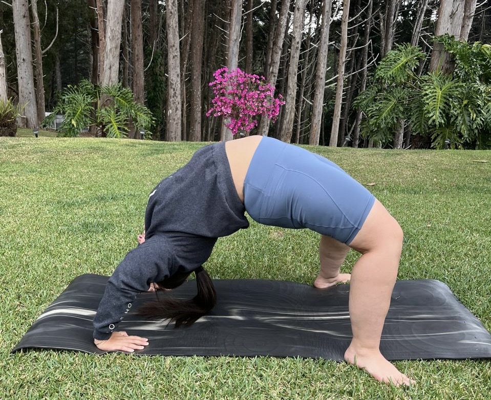 Marce está al aire libre, sobre una colchoneta negra colocada la grama. Está arqueando el cuerpo hacia atrás apoyando manos y pies en el suelo, formando un puente con la espalda elevada. Lleva ropa deportiva y alrededor hay árboles y plantas.
La postura de yoga es conocida como “puente” o “rueda” (Urdhva Dhanurasana).
