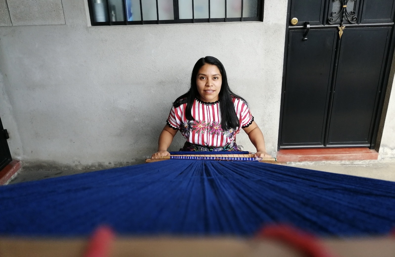 Flory está sentada frente a un telar, trabajando con hilos azules que se extienden de arriba hacia abajo. Lleva una blusa colorida con bordados y tiene el cabello largo y oscuro. Sus manos sostienen el tejido mientras lo va formando con cuidado. El fondo muestra una pared gris y una puerta negra.