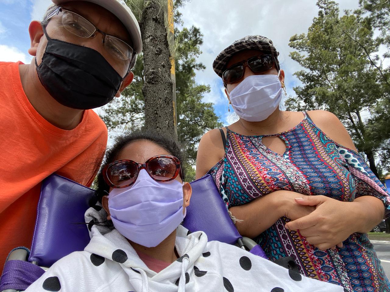 Tres personas están en la calle al aire libre, con árboles y cielo nublado de fondo. En una selfie. Todas llevan mascarillas y lentes de sol. En el centro, está Andrea en su silla de ruedas lleva un suéter blanco con puntos negros. A la izquierda, su papá con camisa naranja y gorra clara. A la derecha, su mamá con gorra y blusa colorida, los dos de pie. 