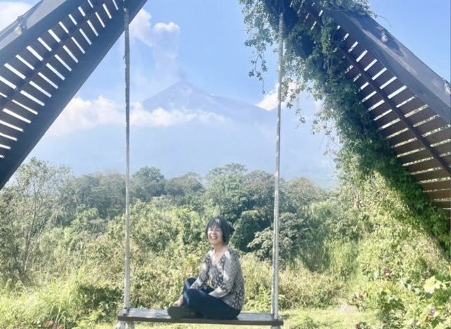 Silvia Quan está sentada en un columpio de madera en medio de un paisaje natural. El columpio forma un triángulo cubierto parcialmente de plantas. Al fondo se ve un volcán bajo un cielo azul con algunas nubes, rodeado de vegetación. Tiene un pantalón de lona azul y una blusa con rombos blancos y negros.